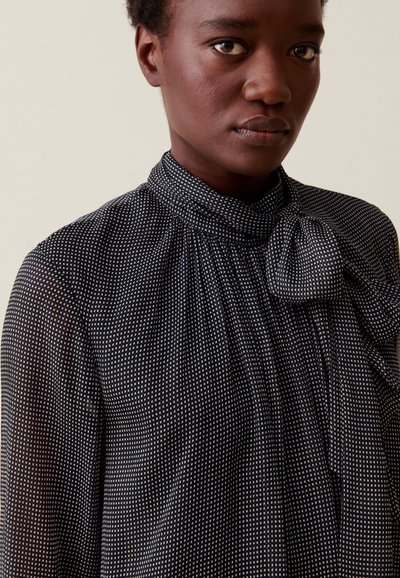 Blusa negra con lunares blancos, cuello alto y detalle de lazo. Tejido transparente con una sutil textura, con acentos fruncidos en los hombros.