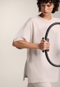 Svijetlosmeđa oversized majica s kratkim rukavima, bočnim prorezima i glatkom teksturom, drži se s dvoje ruku koje čvrsto drže crni vježbački obruč.