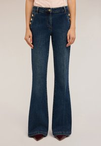 Uitlopende donkerblauwe denim jeans met een hoge taille, voorzien van knoopaccenten op de heup en een gladde textuur. Getailleerde pasvorm tot aan de flares.