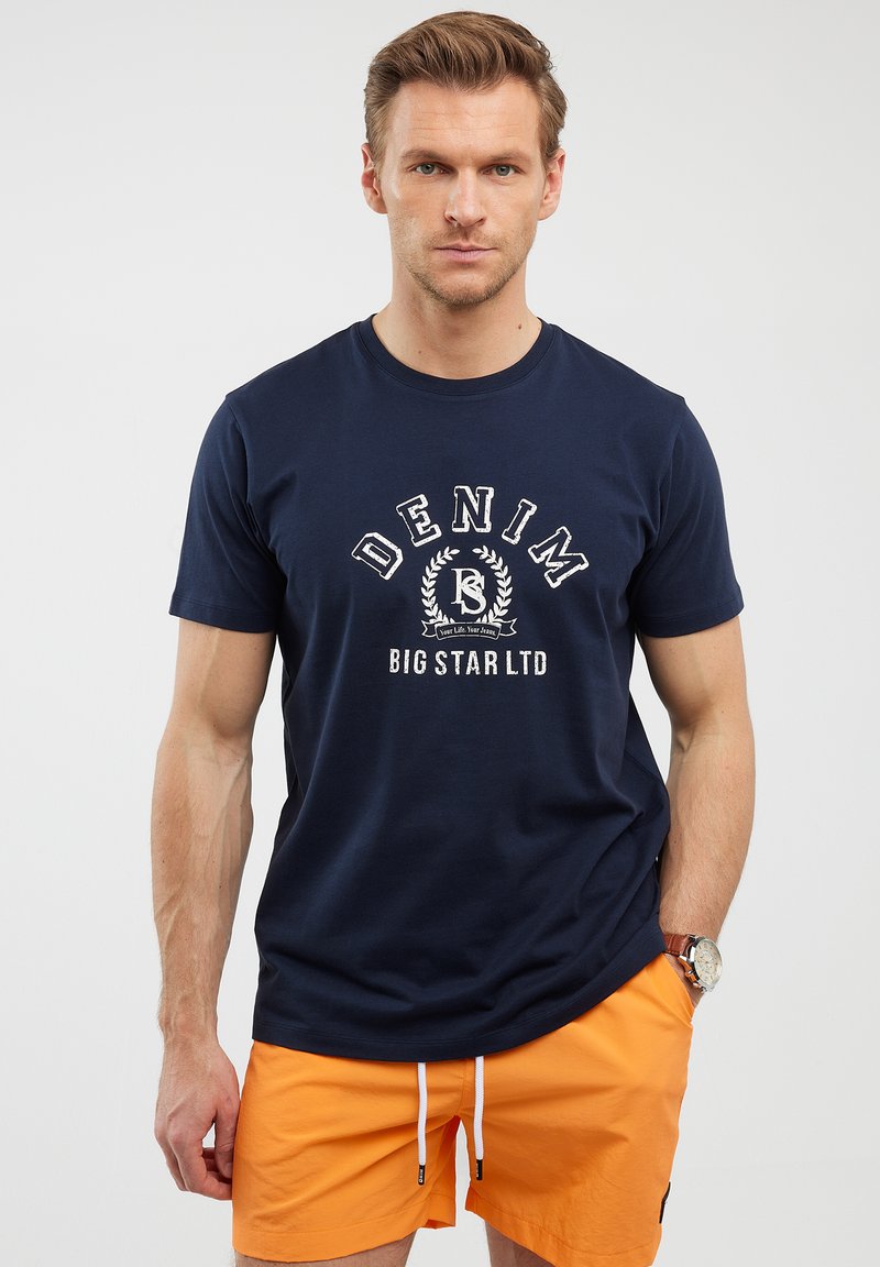 Tamsiai mėlynos spalvos medvilninis marškinėliai su apvalia iškirpte ir spausdintu "DENIM BIG STAR LTD" logotipu, derinami su oranžiniais šortais, laisvas kirpimas.