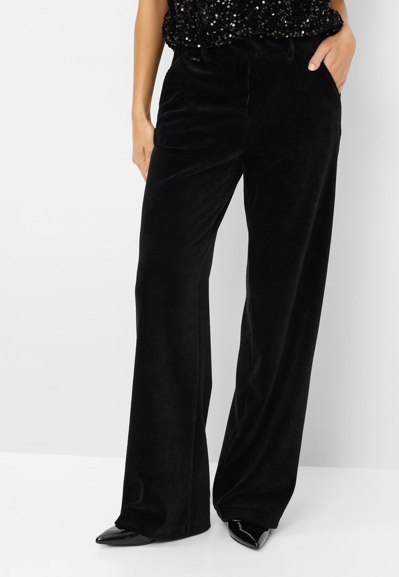 Pantalons en velours côtelé noirs à larges jambes avec une texture douce, présentant des poches latérales et une silhouette droite. Associés à des chaussures noires à bouts pointus.