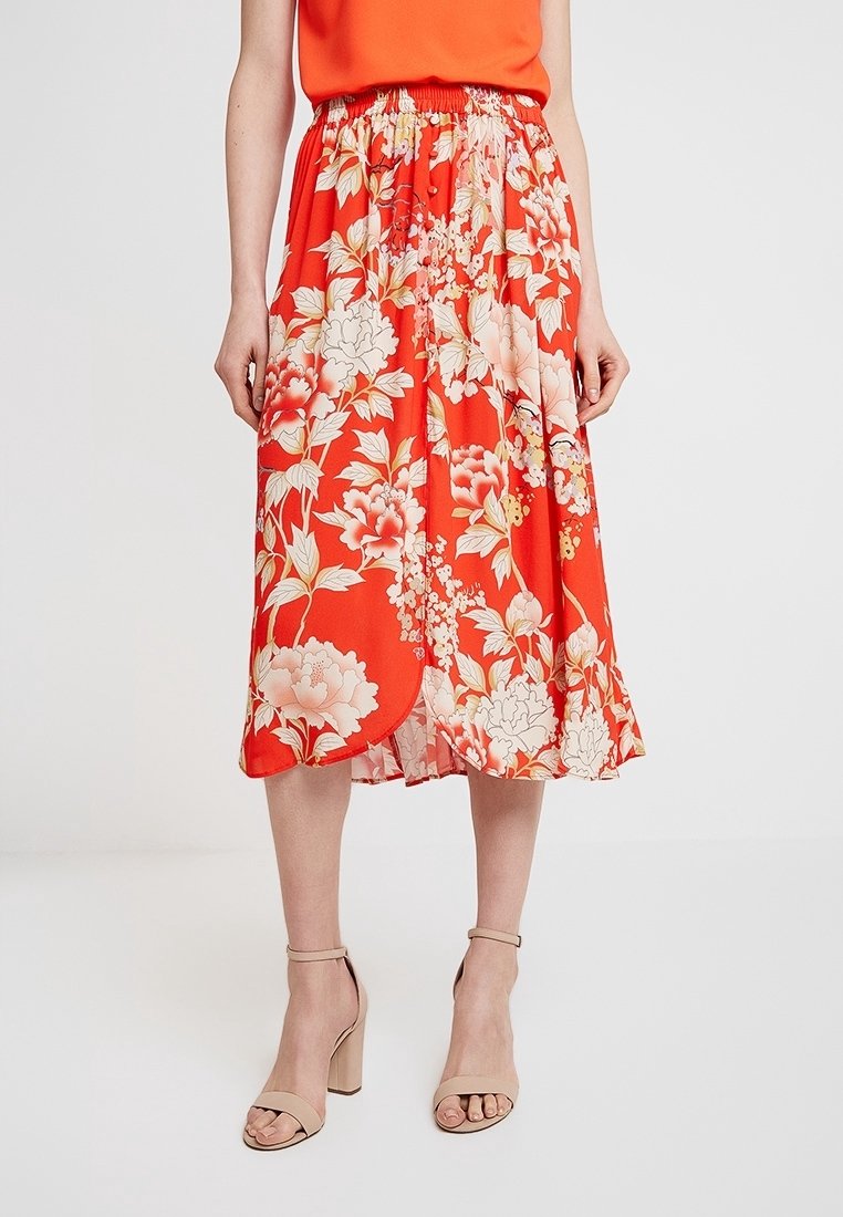 Jupe florale rouge avec une taille froncée, ornée de grandes fleurs crème et roses, silhouette fluide et tissu léger.