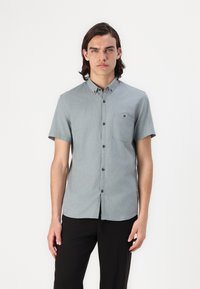 Camisa de botón de manga corta, color gris claro, con un bolsillo en el pecho, textura sutil y botones oscuros en contraste, combinada con pantalones negros.