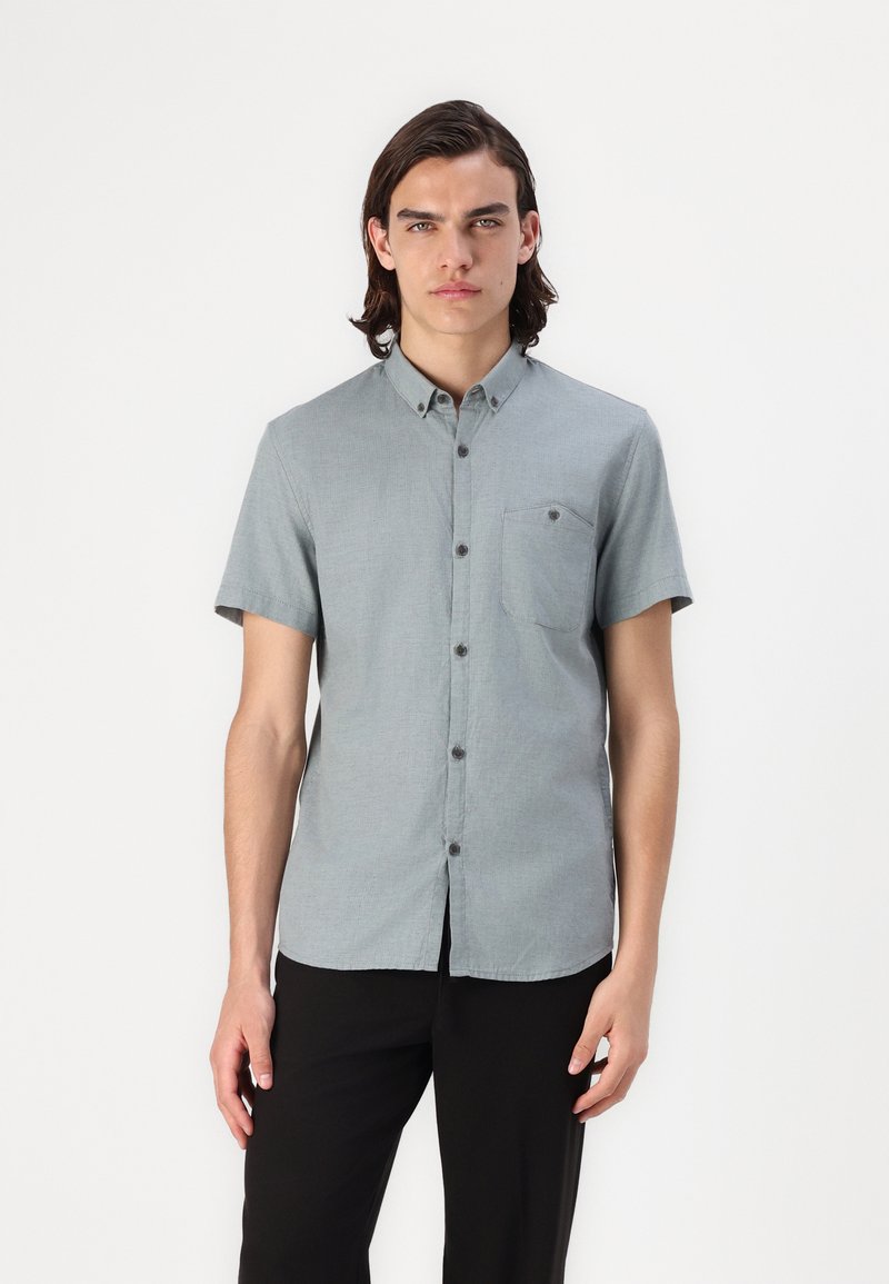 Camisa de botón de manga corta, color gris claro, con un bolsillo en el pecho, textura sutil y botones oscuros en contraste, combinada con pantalones negros.
