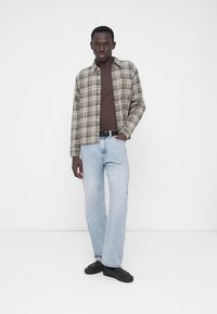 Πουκάμισο καρό με κουμπιά σε μπεζ και σκούρο πράσινο, συνδυασμένο με καφέ μπλούζα και ανοιχτόχρωμα μπλε τζιν. Μαύρα αθλητικά παπούτσια συμπληρώνουν το ντύσιμο.