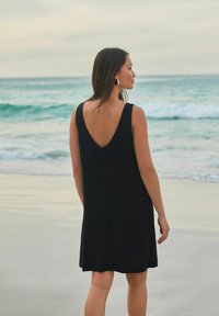 Robe noire sans manches avec un dos en V, faite d'un tissu doux, tombant au-dessus du genou, portée par une personne se tenant sur une plage.