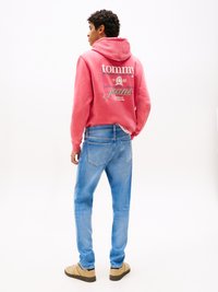 Ružičasti dukserica s logotipom "Tommy Jeans" i grafikom na leđima; uparen s svijetloplavim trapericama i bež obućom. Opušteni, ležerni kroj.