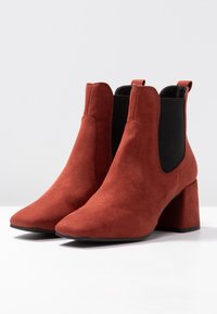 Une paire de bottines en suède couleur rouille avec des talons block et des panneaux élastiques noirs sur les côtés, présentées sur une surface blanche.