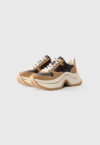 Chunky beige and black sneakers with white laces, thick sculpted soles, and a small round gold logo charm on the side.