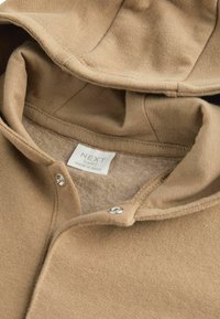 Felpa color beige con un interno morbido eTexture. Presenta chiusure a pressione e un'etichetta con la scritta "NEXT Baby, Made in India." Design e tessuto casual.