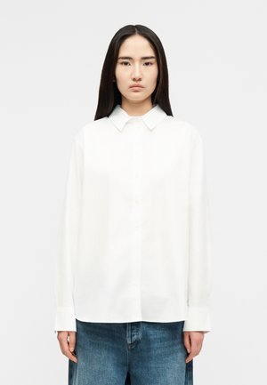 Femme aux longs cheveux foncés porte une chemise blanche ample à boutons et un jean bleu large, debout devant un fond uni clair.