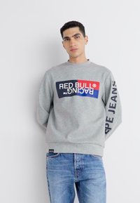 Sivi dukserica s velikim logotipom "RED BULL RACING" u plavoj i crvenoj boji sprijeda, koja uključuje rebrasti ovratnik i manžete. Uske traperice.