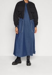 Chaqueta bomber negra con dos bolsillos frontales sobre un vestido largo de mezclilla. El vestido cuenta con dos bolsillos grandes y un dobladillo deshilachado. Las botas de combate negras completan el look.