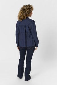 Navyblaues Langarmhemd mit gerafftem Detail auf der Rückseite, kombiniert mit dunklen Schlagjeans. Das Hemd hat einen Kragen und geknöpfte Manschetten.