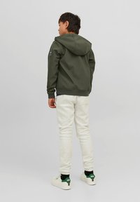 Mørkegrøn hættejakke med en glat overflade, parret med lysegrå sweatpants. Hvide sneakers med grønne detaljer ved hælen.