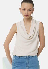 Beżowa bluzka bez rękawów z miękkiego materiału, z drapowanym dekoltem. Połączona z jasnoniebieskimi dżinsami, podkreślającymi luźny krój.