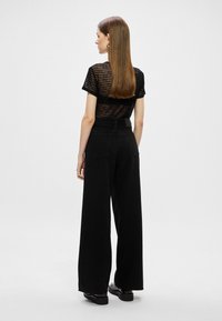 Top preto transparente e texturizado com mangas curtas, combinado com umas calças de ganga pretas de perna larga com dois bolsos traseiros. Sapatos pretos simples completam o visual.