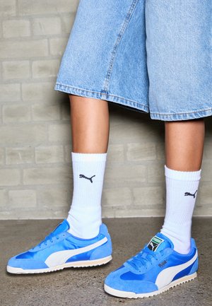 Sneakers blu in suede e rete con accenti bianchi, abbinate a calzini bianchi a coste con un logo nero, indossati con pantaloni corti di jeans chiari.