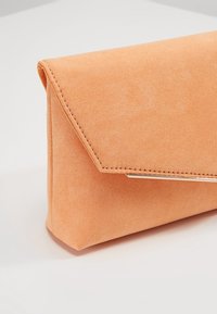 Broskvový semišový clutch s trojúhelníkovým uzávěrem, zlatými akcenty a prošíváním podél okrajů. Obdélníkový tvar.