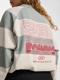 Persoană care poartă un hanorac verde cu dungi albe, cu text roșu „23 Swarm Internationals” și „Serving the game since 1923” pe spate.