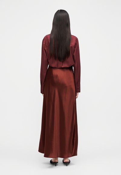 Długa, ciemna sukienka z jedwabiu w kolorze wina, z dopasowaną talią i zwiewnym dołem, z długimi rękawami i zapinana na guziki z tyłu, noszona z czarnymi sandałami na obcasie.