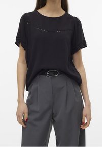 Un blouson noir à manches courtes avec une bande texturée sur la poitrine, associé à un pantalon gris taille haute avec des plis et une ceinture.