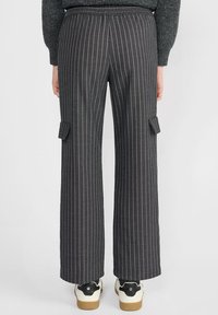 Personne portant un pantalon large à rayures grises foncées avec des poches latérales et des baskets blanches, se tenant devant un fond uni.