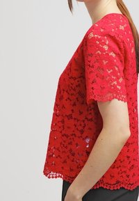 Blusa rossa in pizzo con maniche corte, vestibilità ampia, motivo floreale e orlo scallop. Il materiale è leggero con una finitura testurizzata.