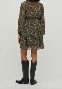 Blommig klänning med långa ärmar, samlad midja och utsvängd kjol, med en svart bakgrund och gula och lila blommor. Svarta stövlar till knäna.