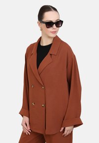 Veste double boutonnage marron avec coupe oversize, grands revers et boutons dorés, portée avec un top noir et des lunettes de soleil modernes.