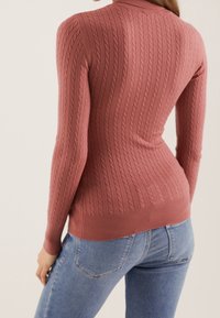 Gerippter Strickpullover in weichem Rosaton, mit hohem Kragen und figurbetontem Schnitt. Dazu kombiniert helle blaue Jeans.