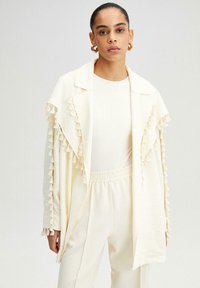 Chaqueta color crema con flecos en los puños, fabricada en un tejido ligero, diseño de abertura frontal, apariencia en capas, combinada con una blusa y pantalones a juego.