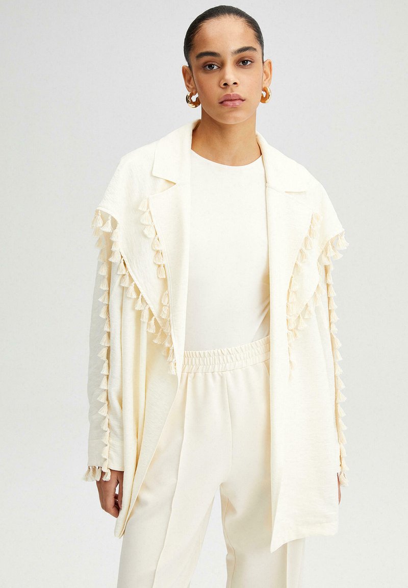 Chaqueta color crema con flecos en los puños, fabricada en un tejido ligero, diseño de abertura frontal, apariencia en capas, combinada con una blusa y pantalones a juego.
