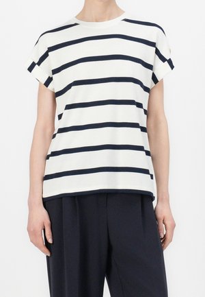 Persona con una camiseta blanca de manga corta con rayas horizontales negras y pantalones negros holgados, de pie con los brazos relajados.