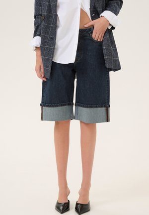 Pantalones cortos de mezclilla azul oscuro con un dobladillo sin terminar, puños de mezclilla azul claro y un detalle de costura minimalista. Combinados con una camisa blanca y un blazer.