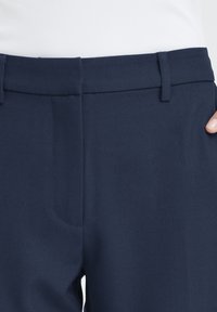Pantalon bleu marine foncé à texture lisse, avec un devant plat, des passants pour ceinture et un détail de poche subtil.