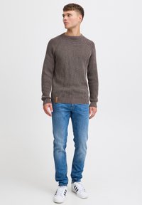 Brązowy sweter o teksturowanym splocie z prążkowanymi mankietami i dołem, w zestawieniu z jasnoniebieskimi dżinsami i białymi trampkami z czarnymi paskami.
