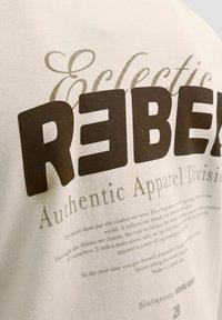 Lähivõte beežist kangast, millel on suur ja tugev tekst "REBEL" ning väiksemad koolitus- ja trükitekstid ehtsa rõivastuse ja eneseväljenduse kohta.