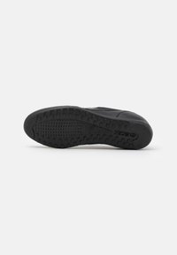 Black shoe with a rubber sole featuring a textured pattern and small circular grips for traction. Sleek design with minimal accents.