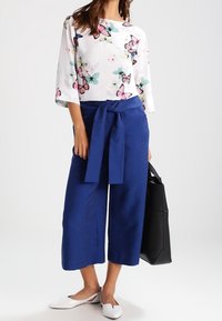 Blouse blanche avec imprimé de papillons multicolores, pantalon large bleu avec ceinture à nouer, ballerines blanches et sac fourre-tout noir.