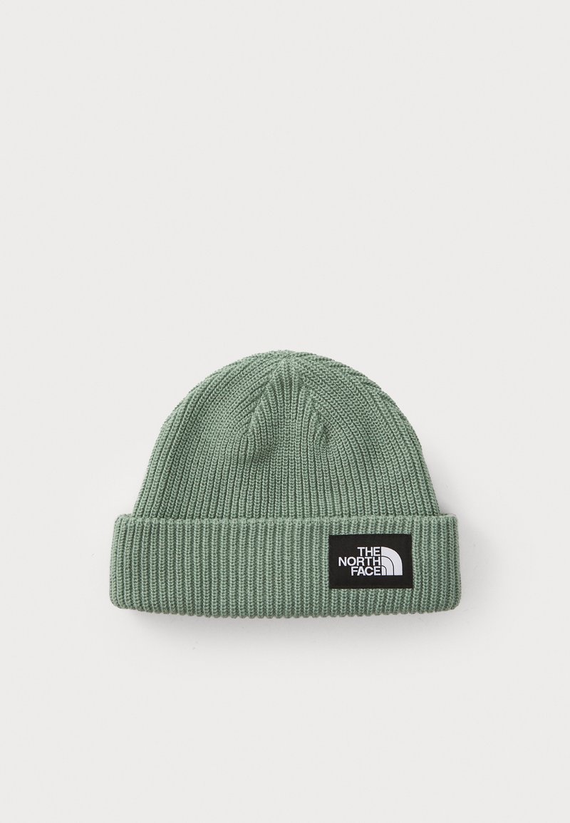 Green knitted beanie with a folded ribbed cuff, featuring a black logo patch with white text. The texture is soft and elastic.