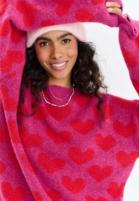 Růžová svetřík s červenými vzory srdcí, jemná textura, široké rukávy a odpovídající růžová čepice. Náhrdelník obsahuje vícebarevné korálky.