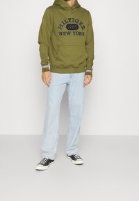 Sudadera verde oliva con logo azul marino, bolsillo canguro y puños acanalados. Jeans rectos de color azul claro y zapatillas negras.