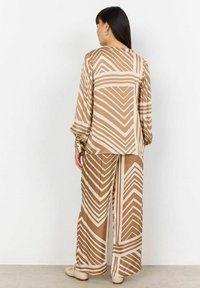 Braunes und cremefarbenes, gemustertes Outfit mit einem langärmeligen Oberteil und gefältelten Schultern sowie weiter geschnittenen Hosen im Zickzackdesign.