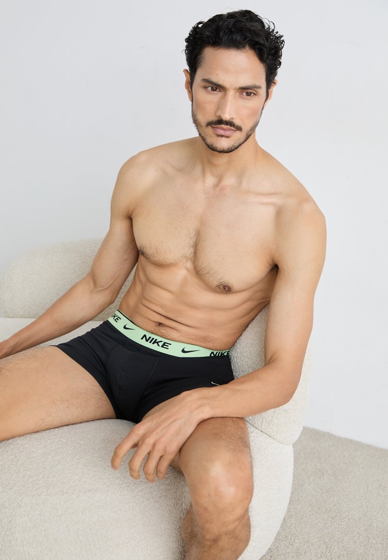 Schwarze Nike-Boxershorts mit einem grünen Bund, auf dem das Markenlogo zu sehen ist. Das Model sitzt in einem hellen, strukturierten Stuhl.