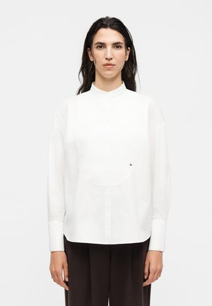 Femme aux longs cheveux foncés portant une chemise blanche boutonnée avec un col montant et un pantalon noir, debout devant un fond uni.