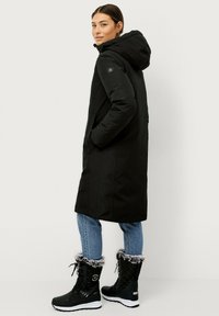 Zwarte gewatteerde parka met capuchon, zijzakken en logo-detail, gecombineerd met zwarte bontgeviseerde winterlaarzen en blauwe jeans. Gladde stoftextuur.