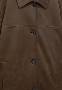 Manteau en suède marron avec un col pointu, cinq boutons et une poche latérale unique. Texture douce avec des accents de boutons foncés contrastants.