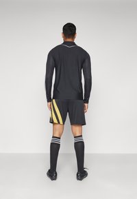 Schwarzes langärmliges Sportshirt mit weißen Akzenten, kombiniert mit schwarzen Shorts, die gelbe diagonale Streifen haben. Schwarze Socken und Schuhe runden das Outfit ab.