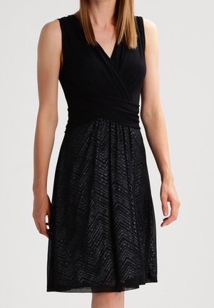 Czarna sukienka bez rękawów z kopertowym dekoltem w serek i wzorzystą spódnicą w linii A, noszona przez stojącą kobietę na jednolitym tle.